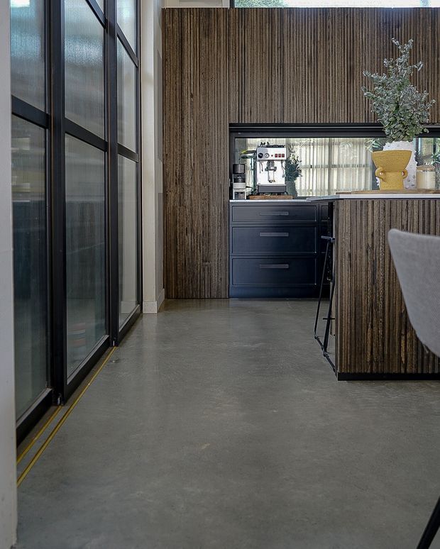 A contemporary kitchen or hospitality-style kitchenette with dark vertically ribbed timber wall and island cladding, matte black cabinetry, a built-in coffee station, and a polished concrete floor. Tall black-framed glazed doors line the left side, while a large framed opening above the cabinetry reflects or reveals a bright adjoining space. The room has a refined industrial character with strong vertical lines and a restrained dark neutral palette.