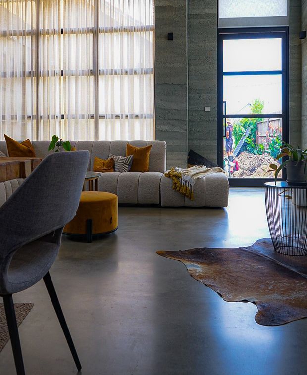 A bright contemporary living room with polished concrete flooring, a large light-grey curved sectional sofa, mustard and ochre accent cushions and ottoman, and a sculptural grey dining chair in the foreground. Floor-to-ceiling sheer window coverings provide filtered daylight, while a tall glazed door opens to a planted exterior. The room includes a black wire-frame side table and an irregular hide rug, with exposed horizontal concrete wall texture adding an industrial character.