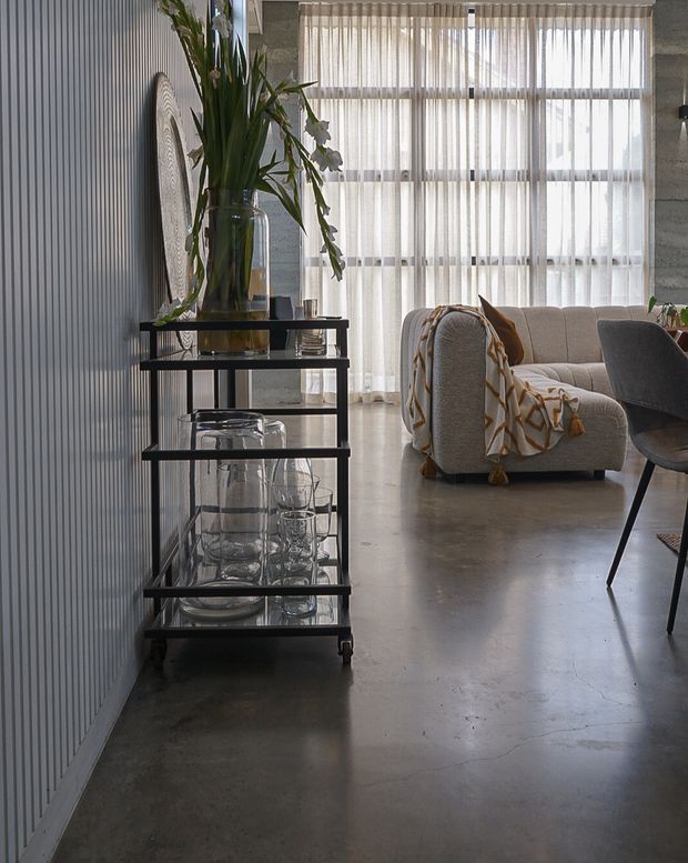 A stylish open-plan living area with polished concrete flooring, a vertically ribbed feature wall, and expansive sheer-curtained windows. A black metal-and-glass bar cart with decorative greenery and glassware sits in the foreground, while a curved light upholstered sofa and dining chair occupy the background. The space has an industrial-contemporary feel with soft, warm furnishings.