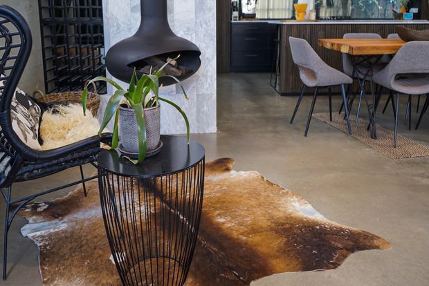 Open-plan interior combining a living area and kitchen/dining zone. A black sculptural suspended fireplace is mounted against a light stone-textured wall, with black wire-frame side tables, a rounded black lounge chair, indoor plants, a timber dining table, upholstered dining chairs, dark cabinetry and polished concrete flooring. A cowhide rug adds a warm brown accent.