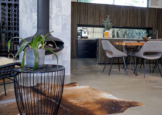 A refined open-plan interior combining a living area and dining-kitchen zone. A sculptural black fireplace sits against a pale stone feature wall, with a black wire-frame side table and potted plant in the foreground. Behind it, dark timber wall panelling frames a compact black kitchen and dining area with a timber table, upholstered chairs, and large glazing. The polished concrete floor and restrained earthy palette create a contemporary, warm-industrial atmosphere.