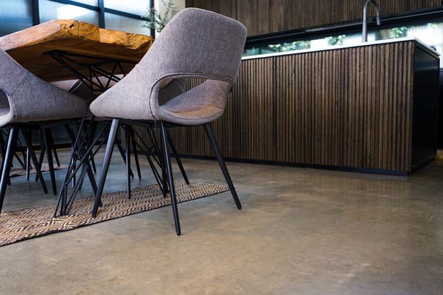 A contemporary open-plan dining and kitchen area featuring a live-edge timber dining table with upholstered grey dining chairs, a patterned rug, polished concrete flooring, and a kitchen island clad in dark vertical timber battens. Large windows and greenery visible beyond provide natural light.