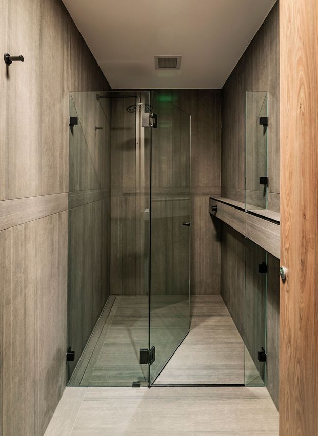 A narrow, contemporary bathroom viewed through an open timber door. The space is dominated by a frameless clear-glass walk-in shower with black hardware, grey stone-look wall and floor finishes, a floating timber-toned vanity with integrated basin, and a large wall mirror. Recessed ceiling ventilation and minimalist black fixtures complete the restrained, dark neutral scheme.