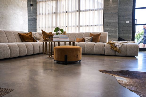 A spacious contemporary living room with a large curved modular upholstered sectional, warm brown cushions, a round leather ottoman, and a slim wood-and-metal coffee table. The room features polished concrete flooring, textured concrete walls, expansive sheer-curtained glazing, and dark-framed windows, creating an industrial yet comfortable atmosphere.