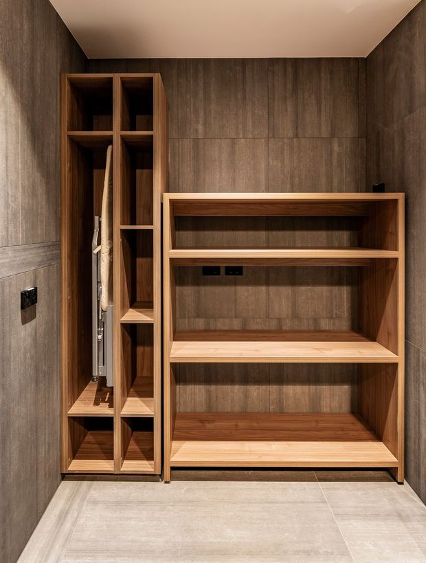 Compact, highly tailored storage room or wardrobe with full-height dark wood-look wall panelling and matching built-in timber joinery. The left unit has narrow vertical compartments and an open hanging section, while the larger right unit provides multiple open shelves and a broad lower shelf. Large pale grey floor tiles and a warm off-white ceiling contrast with the rich brown cabinetry. The image emphasizes efficient custom storage and a clean, refined material palette.