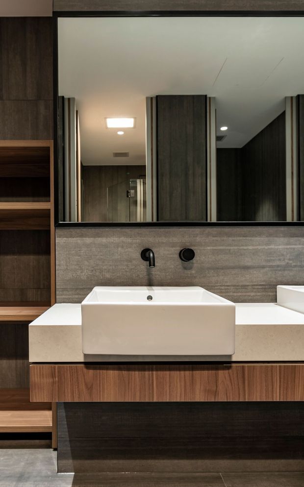Close-up view of a refined contemporary bathroom vanity. A white vessel basin sits on a pale stone or engineered-stone benchtop with a wall-mounted black tap and mixer. Dark timber cabinetry and open shelving frame the vanity, while a large mirror reflects additional dark joinery and a glass shower enclosure. The palette is warm and moody, with layered timber, stone, black fixtures, and recessed lighting.