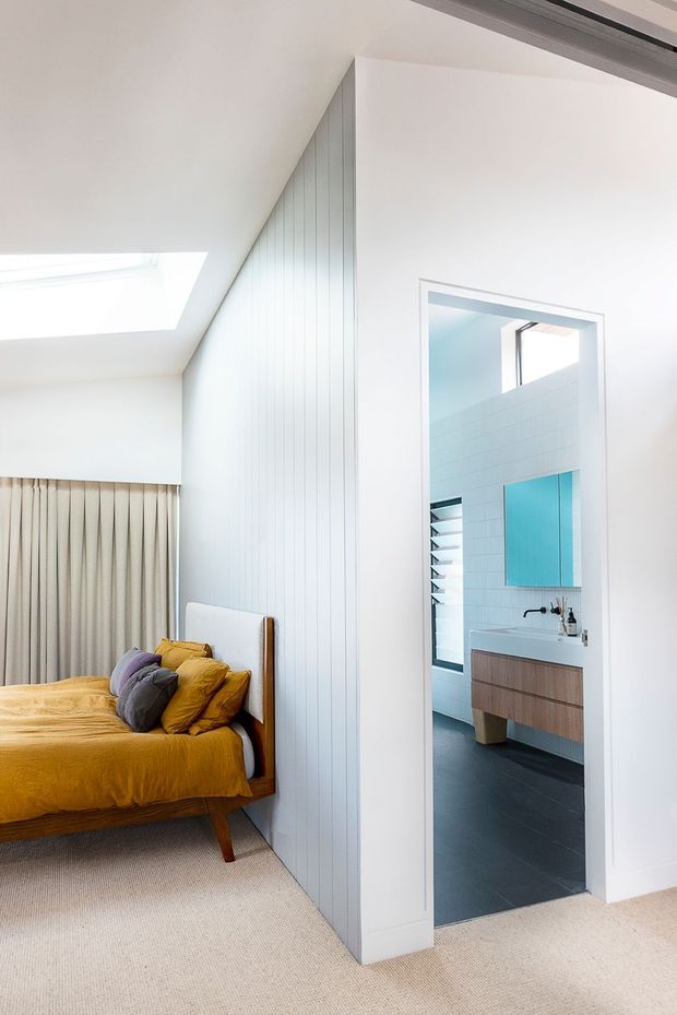 Bright contemporary bedroom with a mustard upholstered bed and layered cushions, beige carpet, full-height curtains, and a white vertical-panel feature wall. A directly connected bathroom is visible through the open doorway, with pale wall tiles, dark floor tiles, a floating timber vanity, and a turquoise mirror or wall panel. A skylight brings additional natural light into the bedroom.