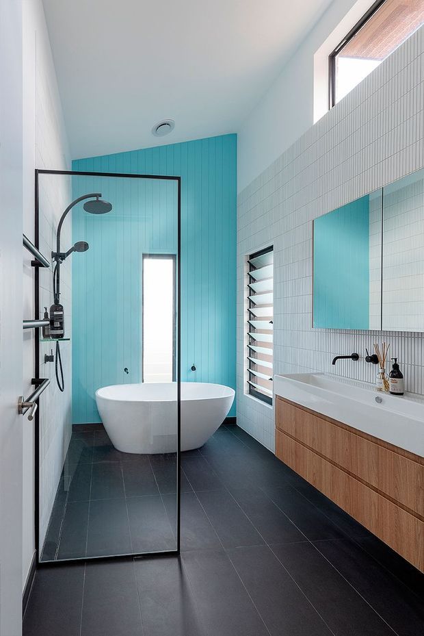 A bright contemporary bathroom with a freestanding white bathtub, walk-in glass shower, wall-mounted timber vanity, and large mirror. The room features a vivid turquoise vertical-panel feature wall, white small-format tiled walls, dark floor tiles, black tapware and shower fittings, and high-level glazing with louvred ventilation windows.