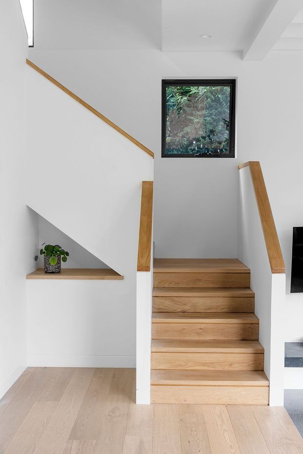 Bright contemporary stairway with a straight timber staircase, white painted walls, integrated timber handrails, and pale timber flooring. A black-framed window brings in views of greenery, while a small recessed ledge holds a potted plant. Clean lines and minimal detailing create an airy, understated interior.