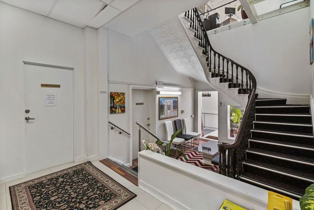 Interior view of a bright institutional or commercial entry hall featuring a sweeping dark timber staircase with ornate turned balusters, white walls, tiled flooring, framed artwork, seating, and patterned rugs. The stair rises to an upper landing and forms the dominant architectural element.