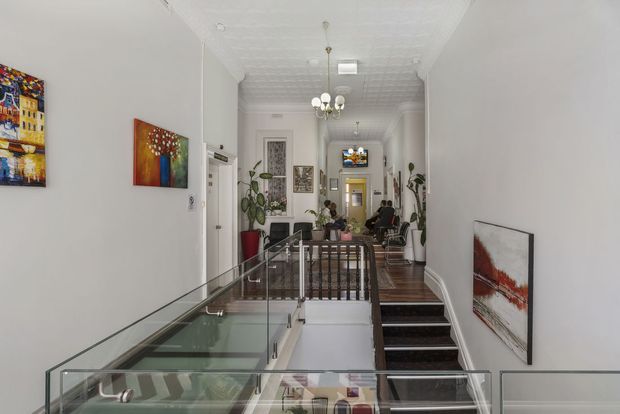 A long, narrow interior hallway and stair landing in a refined period-style home. The space features white walls, an ornate pressed-tin ceiling, decorative ceiling medallion and chandelier, dark timber flooring, a glass balustrade around the stair opening, black stair treads, potted plants, and colourful framed artwork. A sitting area is visible at the far end.