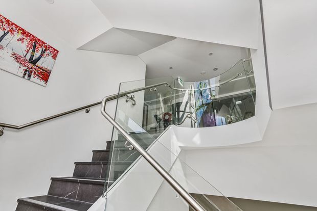 A contemporary interior stairway with dark stone steps, a stainless-steel handrail, and clear glass balustrades. The stairs rise around a sculptural white curved void, with angular ceiling forms, recessed lighting, and reflected artwork visible in the glazing.