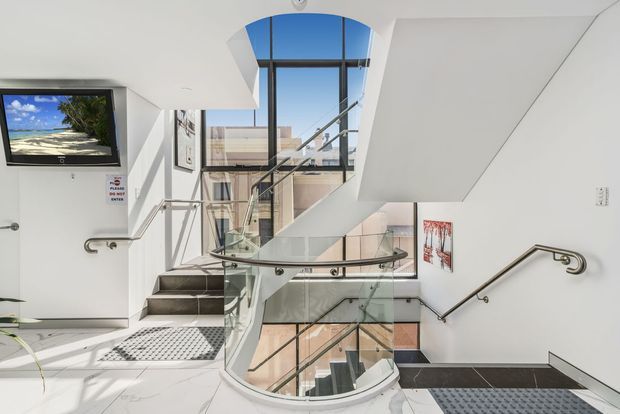 Bright contemporary multi-level stairway with a curved glass balustrade, dark stair treads, stainless-steel handrails, and expansive floor-to-ceiling glazing. A large skylight and open stair void bring in abundant daylight, with white walls and polished light flooring creating a clean, airy atmosphere.