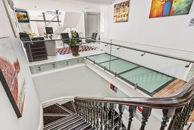 Bright multi-level commercial interior viewed from a staircase, featuring a polished dark timber balustrade, glass bridge and stair-side balustrades with metal fittings, white walls, artwork, office desks, and a patterned carpeted stairway. The open circulation area includes a glazed floor section and a small office/reception zone.