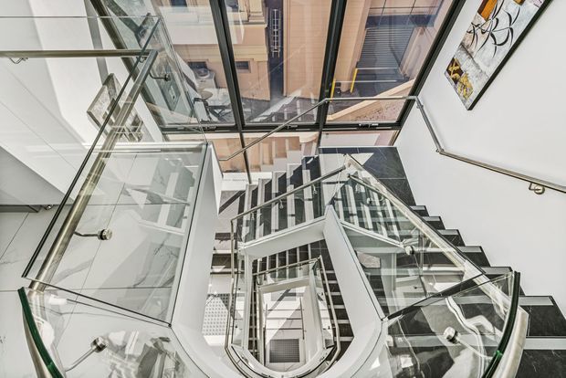 Looking upward through a multi-level interior stairwell with a sculptural curved staircase, dark stone stair treads, white structural walls, extensive glass balustrades, and a glazed roof or ceiling. The space is bright and highly architectural, with surrounding upper-level balconies and framed artwork visible.