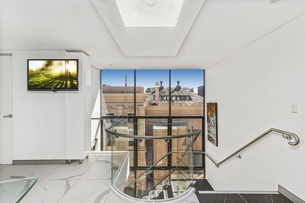 Bright contemporary stairway and landing with a curved glass balustrade, stainless-steel handrails, large black-framed windows, white walls, and a skylight. The view looks across to neighbouring urban buildings, while a wall-mounted television and artwork are visible on the landing.