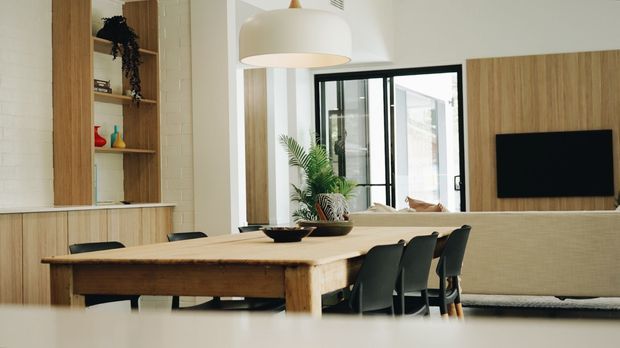 Open-plan contemporary dining and living area with a long natural timber dining table, dark dining chairs, a large cream pendant light, and built-in timber shelving and television joinery. White painted brick and plaster walls, black-framed glazing, and a neutral upholstered sofa create a warm, refined interior.
