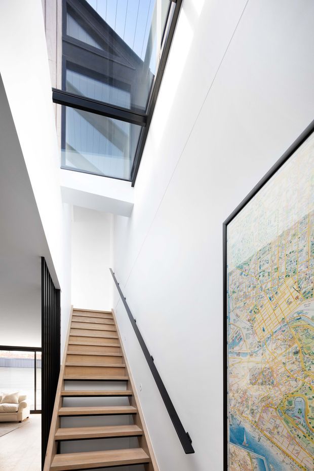 A bright, minimalist interior stairway with light timber open-riser stairs, a slim black handrail, white walls, and a large black-framed skylight or overhead glazing. A framed map hangs on the right wall, while a partially visible living area appears beyond a dark vertical partition at the lower left.