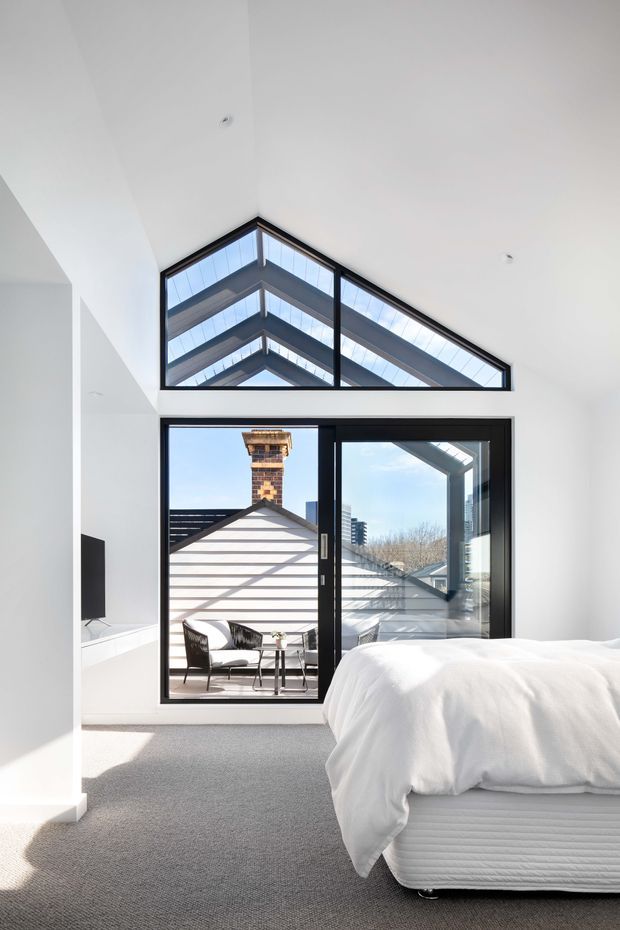Bright contemporary bedroom with a high pitched glazed gable and black-framed floor-to-ceiling sliding doors opening to a small balcony or terrace. The room has white walls, grey carpet, a white upholstered bed, and a minimal wall-mounted television. Strong daylight enters through the glazing, with neighbouring pitched roofs and brick chimney visible outside.