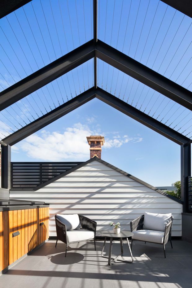 A bright rooftop terrace or balcony with a contemporary black steel pergola structure and overhead tensioned cable canopy. The terrace includes two woven lounge chairs with white cushions, a small round table, and a warm timber-clad built-in cabinet or planter at the side. A white weatherboard gable roof and brick chimney are visible beyond the dark balustrade, with strong linear shadows cast across the grey terrace floor.