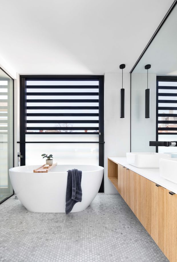 A bright contemporary bathroom featuring a freestanding white bathtub, double vanity with a timber-grain wall-mounted cabinet, white vessel basins, black tapware, and slim black pendant lights. The room has pale tiled walls, a small-format grey mosaic floor, large black-framed windows with striped roller blinds, and a full-height mirror reflecting the vanity and window.