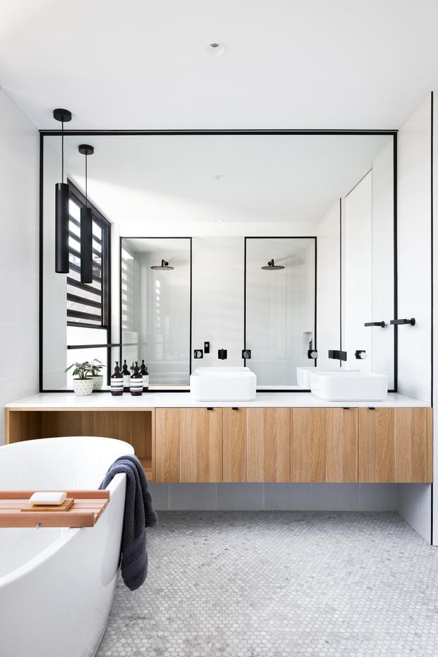 Bright contemporary bathroom with a double floating timber vanity, two white vessel basins, large frameless and black-framed mirrors, dual showers, black fixtures, a freestanding bathtub, and small light-grey mosaic floor tiles. White walls and ceiling create a clean, high-contrast interior, with black pendant lights and warm timber cabinetry adding definition.