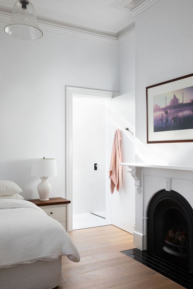 Bright, refined bedroom with a partially visible bed, bedside table and lamp, ornate white ceiling cornices and wall trim, a classic fireplace with black surround and tiled hearth, and an open doorway leading to a white-tiled bathroom. A framed architectural artwork and soft blush towel add restrained colour.