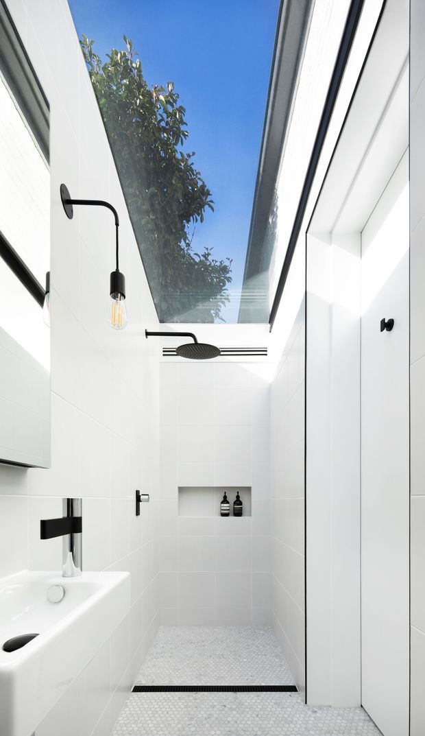 Bright, narrow contemporary bathroom with white tiled walls and a small white hexagonal mosaic floor. The shower area is enclosed by clear glass and topped by a large skylight, with a black ceiling-mounted rain shower, black wall fixtures, a wall niche with dark bottles, and a white vanity basin with a chrome tap. Minimal black accents create a crisp, high-contrast look.