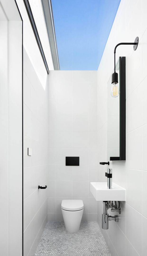 A narrow, bright contemporary bathroom with white tiled walls, a skylight, and a small windowless-style layout. The room features a wall-hung toilet, compact white wall-mounted basin with exposed chrome plumbing, a tall black-framed mirror, and black minimalist wall lighting and fixtures. The floor is finished with small light-grey mosaic tiles, creating a clean high-contrast monochrome scheme.