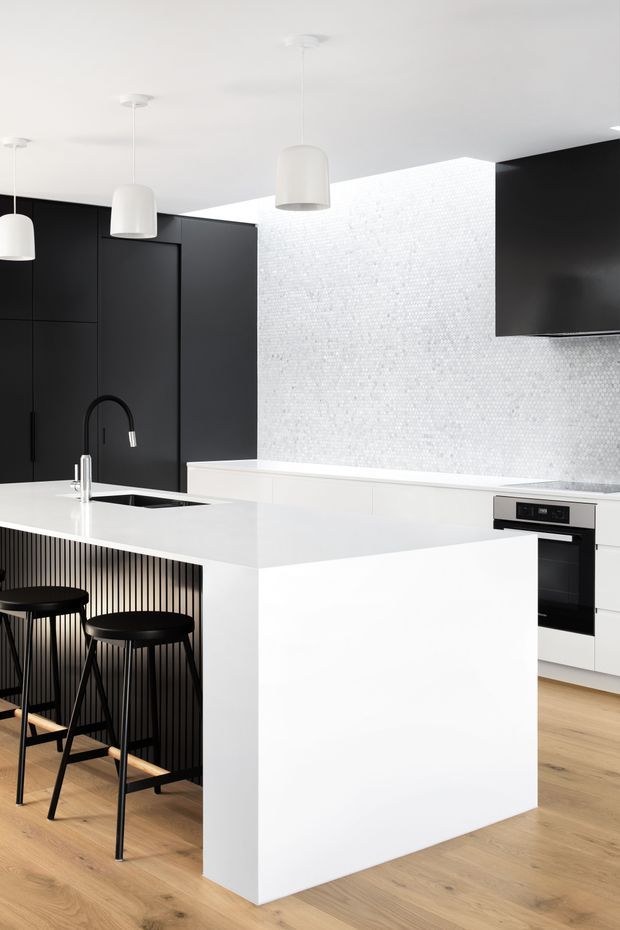 A bright, contemporary kitchen centred on a large white island with an integrated sink and black gooseneck tap. The island includes seating for two at a black slatted peninsula, with black stools. Full-height matte black cabinetry contrasts with a white textured mosaic-tile backsplash, white benchtops, integrated white cabinetry and a built-in oven. White pendant lights hang from a smooth white ceiling, while light timber flooring adds warmth.