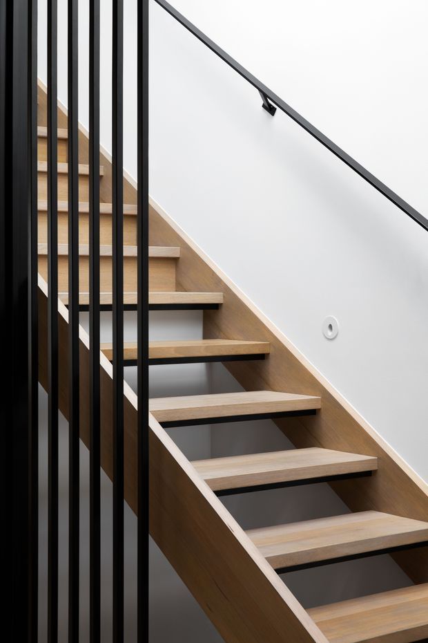 A close-up architectural photograph of a modern open-riser staircase. Broad light timber treads are supported by dark structural elements and framed by a vertical black metal balustrade on the left. A slim dark handrail runs diagonally along a clean white wall, with a small circular wall fitting visible. The composition is minimalist and geometric, emphasizing the contrast between warm timber, black metal, and white plaster.