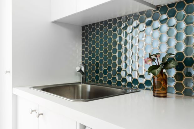 Close-up of a compact, bright kitchen or kitchenette showing white cabinetry, a stainless-steel undermount sink, a white benchtop, and a striking teal hexagonal tile splashback with metallic grout. A small potted orchid sits on the counter.