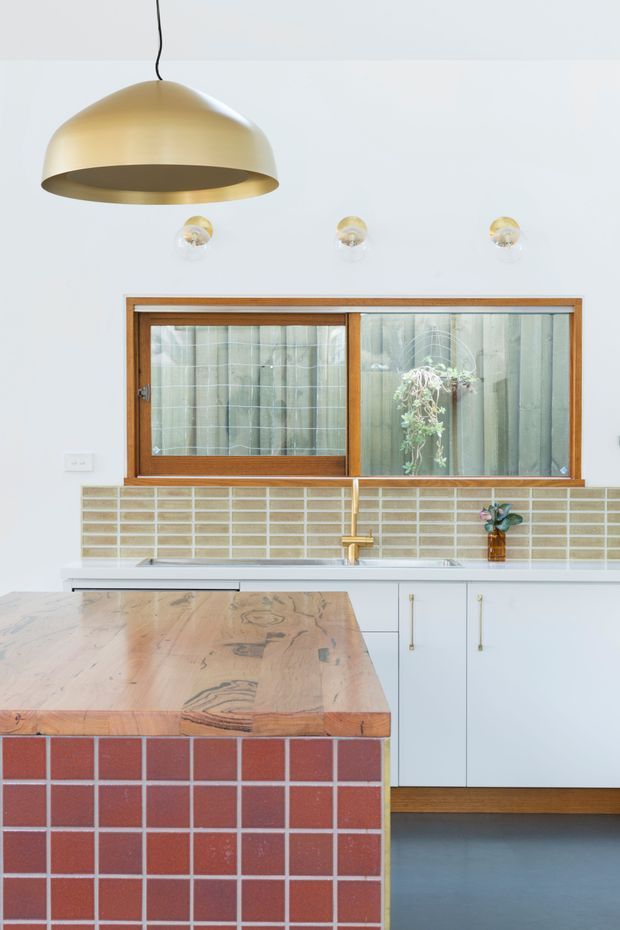 Bright kitchen interior featuring a large timber island with a red ceramic-tile base and timber benchtop, white cabinetry, a pale beige tiled splashback, and a wood-framed sliding window. A large brass pendant light and three small globe lights hang above the island, creating a warm focal point against the white walls.