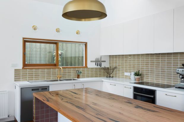 Bright contemporary kitchen with white flat-panel cabinetry, warm timber-framed window, horizontal beige tiled splashback, white benchtops, and a large natural timber island with a dark tiled base. A brass pendant light and brass tap add warm metallic accents.