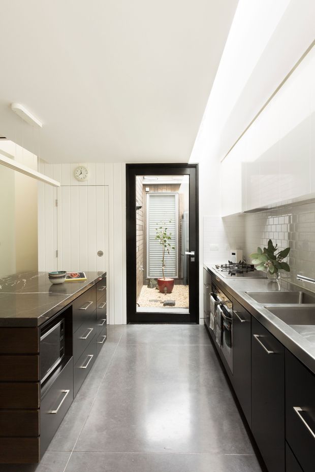 A narrow, galley-style kitchen with dark timber cabinetry, stainless-steel appliances and benchtops, large grey floor tiles, and glossy white overhead cabinets. A white subway-tile splashback runs along the cooking and sink wall. The room is brightly lit by linear ceiling lighting and daylight through a black-framed glazed door leading to a small courtyard.