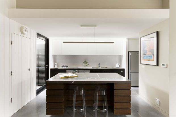 Bright contemporary kitchen viewed straight on, centred on a large dark timber island with waterfall-style pale stone benchtop and seating for three on clear stools. White vertical wall panelling, dark-framed glazing, stainless-steel appliances, a pale tiled or stone splashback, and streamlined pendant lighting create a clean, refined interior.