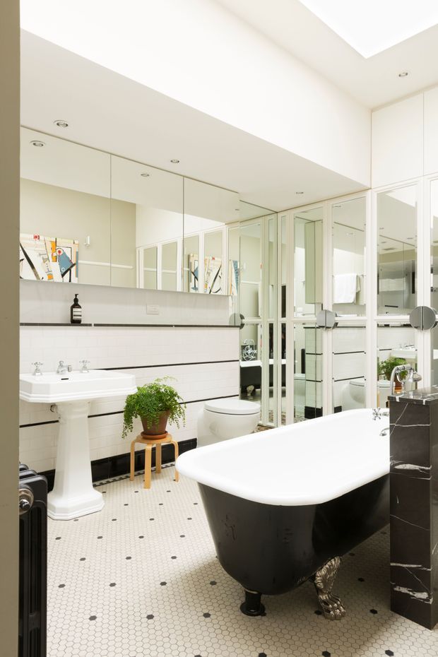 Bright, elegant bathroom featuring a freestanding clawfoot bathtub with a dark exterior, white pedestal basin, toilet, mirrored wall cabinets, white subway wall tiles with dark grout, and a small-pattern hexagonal tiled floor. A potted plant and framed artwork add decorative touches.