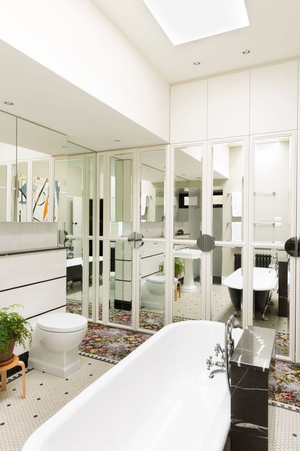 Bright, spacious bathroom featuring a freestanding white bathtub, toilet, tiled vanity, extensive mirrored cabinetry and glass-panelled doors. The room has white walls and cabinetry, small white mosaic floor tiles with dark dot accents, recessed lighting, and a patterned floral rug. Mirrors create multiple reflections of the bathroom and its fixtures.