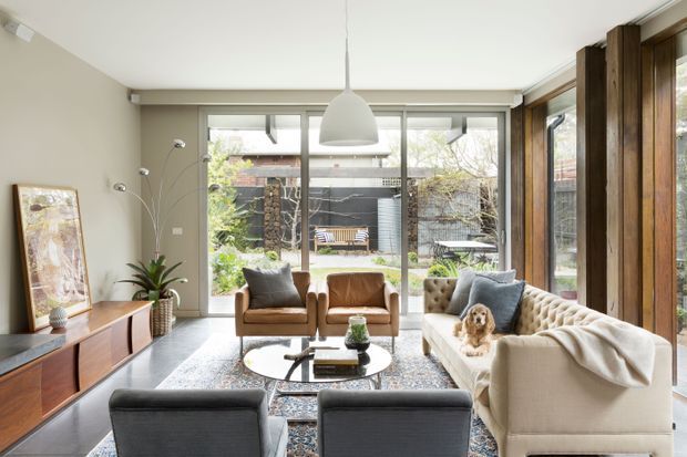 A bright, contemporary living room with floor-to-ceiling glazing opening toward a landscaped courtyard. The room features a cream sofa, two tan leather armchairs, dark upholstered occasional chairs, a round coffee table, patterned rug, timber media console, indoor plants, and a large white pendant light. Vertical dark timber framing and extensive windows create a strong connection to the garden.