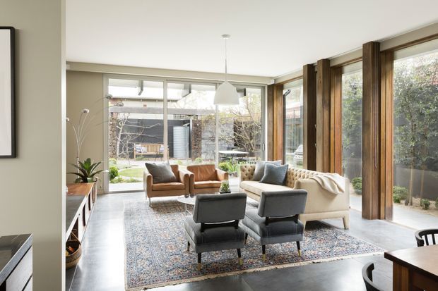 Bright, refined living room with floor-to-ceiling glazing and substantial timber-framed windows overlooking a landscaped courtyard. The room features a large patterned rug, cream sofa, upholstered armchairs, dark lounge chairs, a suspended white pendant, indoor plants, and a dining area partially visible at the right.