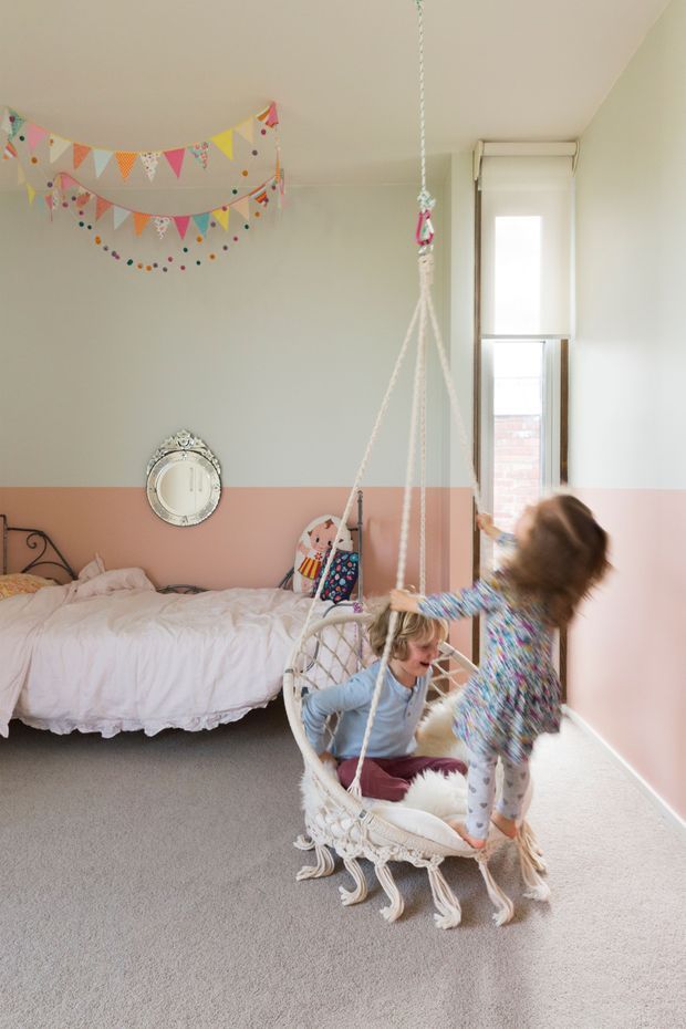 A bright children's bedroom with pale sage-green and blush-pink two-tone walls, a low bed with white bedding, colourful bunting and garlands, a window with a roller blind, and a hanging macramé swing chair occupied by children. The room has a soft, playful, feminine style and carpeted flooring.