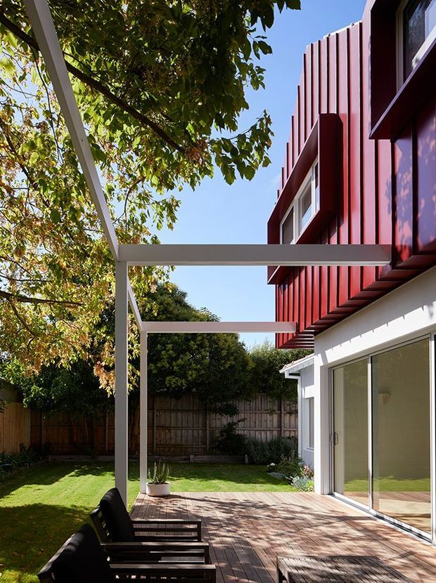 Sunlit rear exterior of a contemporary multi-level home, viewed from a timber deck or patio. The building combines white lower-level walls and large sliding glass doors with a distinctive deep red vertically ribbed upper facade. A minimalist white pergola frame extends over the deck, which includes dark outdoor lounge chairs, with a private fenced garden and mature trees beyond.