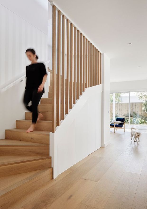 Bright contemporary residential hallway and stairway with pale timber flooring and open-riser-style timber stairs. A white built-in wall runs beneath the staircase, while evenly spaced vertical timber balusters create a sculptural screen along the upper stair edge. The space opens into a light-filled living area with large glazing, and a blurred person and dog are present.
