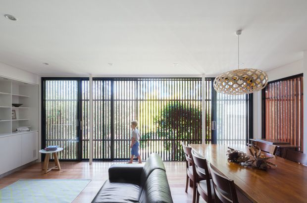 Bright open-plan interior combining a dining area and living room. The space features a long timber dining table with dark upholstered chairs, a black leather sofa, built-in white shelving and cabinetry, and extensive floor-to-ceiling glazing fitted with vertical blinds and dark timber screening. A woven pendant light hangs above the dining table, with greenery visible outside.