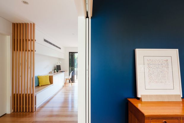 Interior view from an entry or hallway toward a bright open living area, with a vertical timber slat screen, white built-in cabinetry, timber flooring, and a deep blue feature wall. A framed artwork rests on a warm timber console in the foreground.