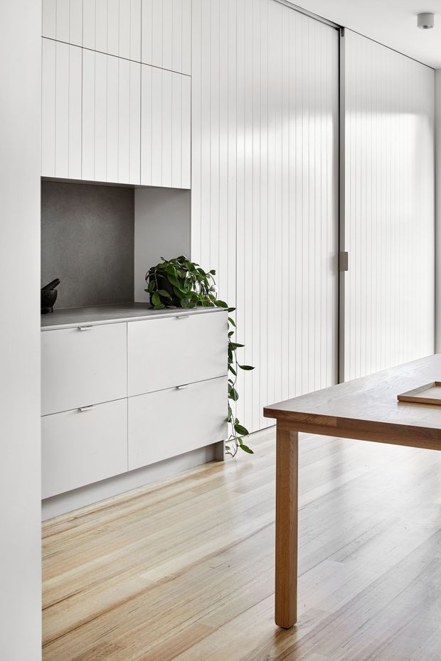 Bright minimalist interior featuring a white built-in storage unit with vertically grooved doors and drawers, a dark grey recessed niche, pale timber flooring, and a simple solid timber dining table. A large vertically grooved sliding or folding door occupies the right side of the room, with a small indoor plant adding greenery.