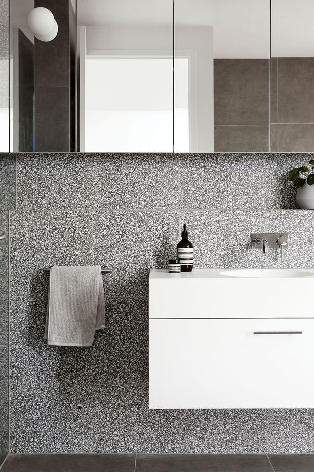 A compact contemporary bathroom featuring a wall-mounted white vanity with integrated basin, chrome wall-mounted tapware, a large mirror, and a grey-and-white terrazzo finish covering the walls. Dark grey floor tiles, a simple towel rail, spherical wall light, and minimal accessories complete the monochrome scheme.