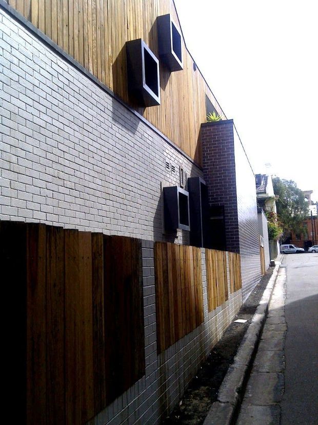 Street-level view of a contemporary multi-level building facade in a narrow urban lane. The lower facade is clad in pale grey brick, while the upper portion uses vertical timber boarding. Dark projecting box windows and a dark brick vertical volume add strong geometric accents. Timber slat fencing and a narrow concrete-paved footpath line the building.