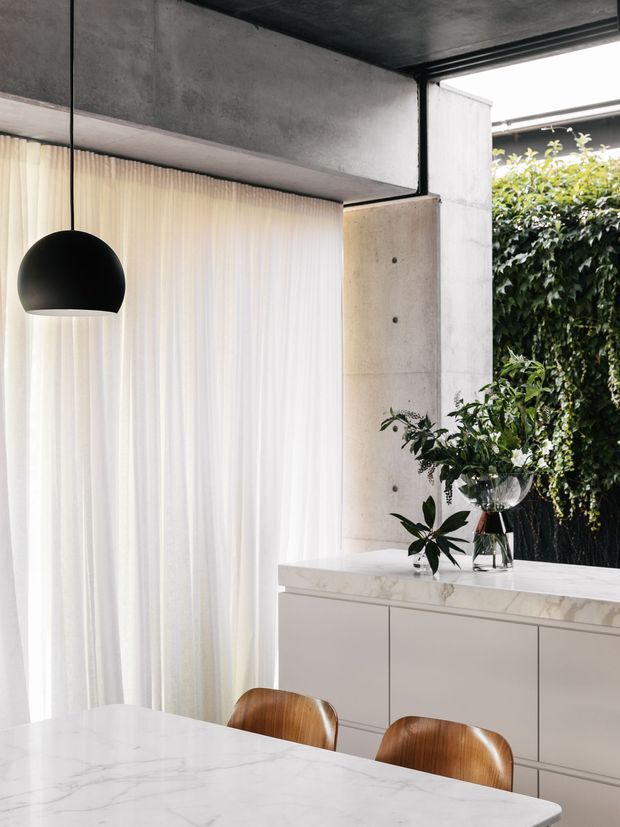 A refined contemporary dining area with a white marble-look dining table and matching built-in sideboard, warm curved timber dining chairs, sheer floor-to-ceiling curtains, and a black suspended dome pendant. Exposed concrete walls and ceiling contrast with the pale cabinetry, while lush greenery is visible through the opening at the rear.