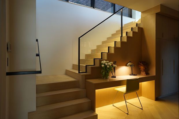 A warm, contemporary stairway with broad timber steps, a slim black-framed glass balustrade, and integrated timber cabinetry beneath the staircase. The under-stair joinery forms a compact illuminated desk nook with a chair, desk lamp, and decorative vase. Clerestory glazing above brings in natural light.